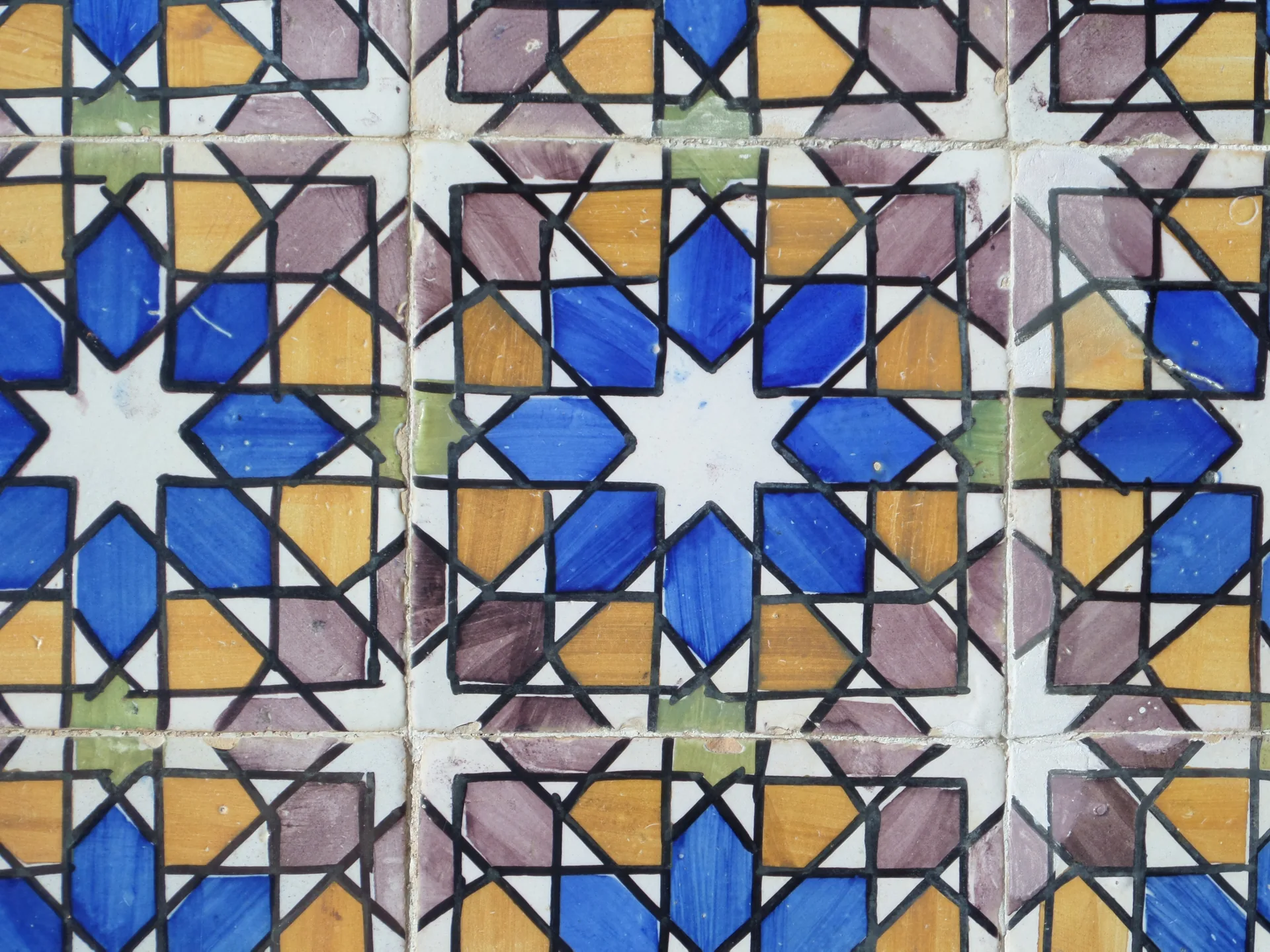 Close-up of decorative ceramic tiles showing a repeating structural pattern in blue, gold, purple, green, white, and black, each tile consistent with the ones around it.